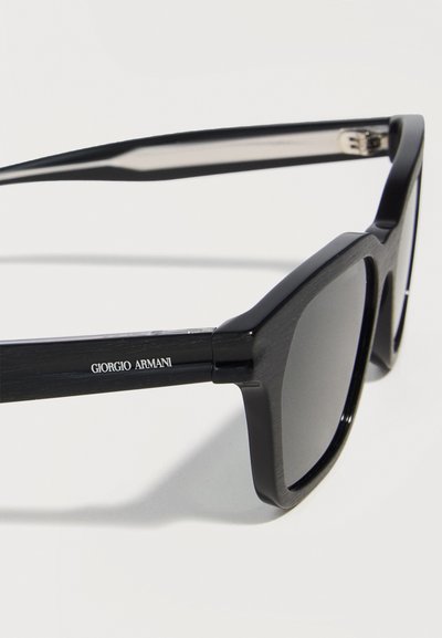 Μαύρα ορθογώνια γυαλιά ηλίου με παχιά σκελετά και λογότυπο "Giorgio Armani" στο σκέλος, εμφανίζονται σε απλό φωτεινό φόντο.