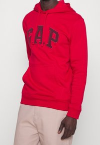 Felpa con cappuccio rosso caratterizzata da un grande logo "G.A.P." grigio scuro. Realizzata in un tessuto morbido, con tasca a marsupio e polsini a costine.