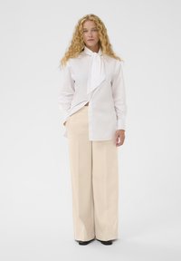 Chemise blanche à manches longues avec une cravate, assortie à un pantalon large couleur crème, présentant une texture lisse et une coupe décontractée.