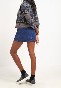 Femme portant une veste à motifs, une mini-jupe en denim bleu, des chaussettes noires et des baskets grises à enfiler, se tenant les mains derrière le dos.