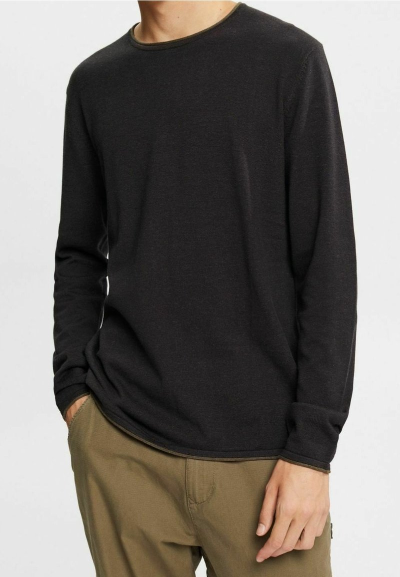 Homme portant une chemise noire unie à manches longues avec un pantalon vert olive, debout avec une main dans la poche, sur un fond clair uni.