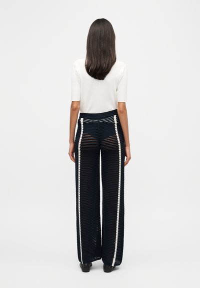 Μαύρο πλεκτό παντελόνι με φαρδιά παντελόνια και λευκές ρίγες στο πλάι. Το ύφασμα έχει υφή με ημιδιαφανές σχέδιο. Το μοντέλο φοράει λευκό τοπ.