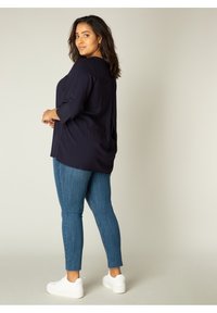 Navyblauwe blouse met driekwartmouwen, textuurstof, losse pasvorm; gecombineerd met blauwe skinny jeans en witte sneakers.