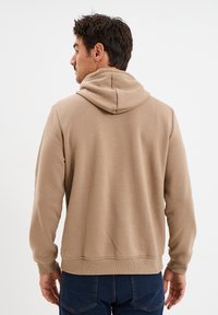 Beige Kapuzenpullover mit weichem Material, gerippten Bündchen und Saum sowie einer Kängurutasche, die auf dem Rücken eines Modells vor einem schlichten Hintergrund positioniert ist.