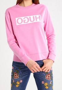Sudadera rosa de manga larga y cuello redondeado, con un logo rectangular blanco de "HUGO" en la parte frontal. Combinada con jeans azules bordados.