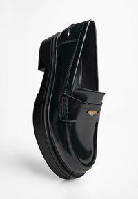 Zwarte gepatineerde leren loafer met een flatte zool, voorzien van een decoratieve band en een klein logo-accent nabij het teengebied.