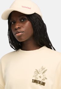 Beige sweatshirt med blommig grafik och texten "Timberland EST. 1973" i brunt. Mjuk tyg och rundad halslinning. Vit keps med rosa logotyp.