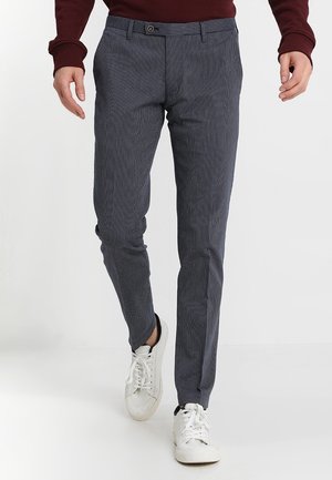 Man loopt en draagt nauwsluitende donkergrijze broek met subtiel patroon en witte sneakers tegen een effen witte achtergrond.