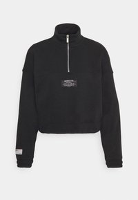 Svart kortärmad sweatshirt med hög krage, fyrakantsdragkedja framtill, texturerat tyg och en etikett på bröstet. Ribbstickade ärmslut har en färgad accent.