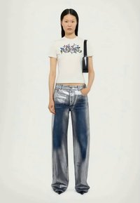 Balta T-maikė su gėlių dizainu, derinama su metalizuotais sidabriniais džinsais, turinčiais laisvą siluetą ir subtilų mėlyną gradientą. Juoda rankinė.