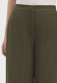 Pantalones de pierna ancha en verde oscuro, fabricados en un tejido texturizado con un sutil patrón de arrugas. Cuentan con una cinturilla recta y una abertura frontal.