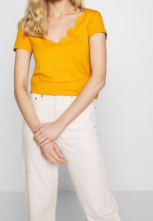 Femme portant un haut à col festonné jaune moutarde à manches courtes et un pantalon blanc taille haute, les mains doucement croisées devant.