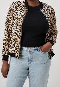 Bomberjacke mit Leopardenmuster, mit einem Reißverschluss vorne, gerippten Manschetten und Saum. Getragen über einem schwarzen Oberteil mit hellblauen, hochgeschnittenen Jeans.