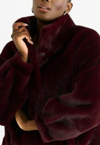 Person wearing a dark burgundy faux fur coat, holding the collar with one hand, wearing hoop earrings, against a plain white background.