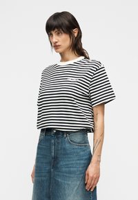 Ριγέ μαύρο-λευκό T-shirt με στρογγυλή λαιμόκοψη και κοντά μανίκια, φορεμένο με μπλε τζιν φούστα. Διαθέτει μικρό λογότυπο στο στήθος.