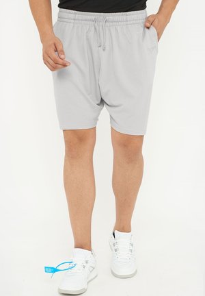 Uomo che indossa pantaloncini grigio chiaro con vita elasticizzata e scarpe da ginnastica bianche, in piedi con una mano in tasca su sfondo bianco.