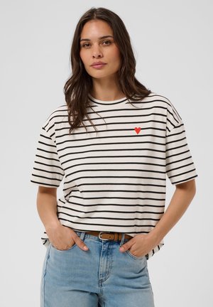 Mujer con cabello largo y castaño lleva una camisa blanca con rayas negras y un pequeño corazón rojo, combinada con vaqueros azul claro y cinturón marrón.
