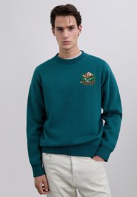 Joven con sudadera color verde azulado con bordado de montaña y árboles, combinado con pantalones beige claro, posando frente a un fondo liso.