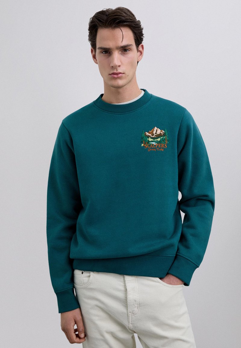 Joven con sudadera color verde azulado con bordado de montaña y árboles, combinado con pantalones beige claro, posando frente a un fondo liso.