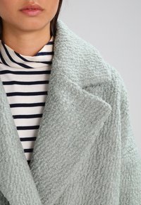 Cappotto testurizzato azzurro chiaro con un ampio colletto, indossato sopra un dolcevita a righe bianche e nere, realizzato in morbido tessuto peluche.