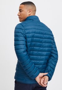 Blaue gepolsterte Jacke mit Stehkragen, horizontalen Steppnähten und glatter Textur. Die Ärmel sind enganliegend und leicht tailliert.