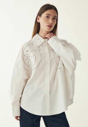 Biela bavlnená košeľa s detailnou vyrezávanou výšivkou na ramenách a klasickým golierom. Má zapínanie na gombíky vpredu a voľný strih.