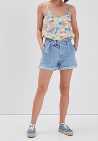 Camisole florale en jaune, bleu et rose. Short en jean léger avec revers roulés et ceinture. Chaussures bleues avec lacets blancs et accents en jute.