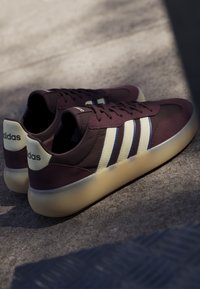 Paire de baskets Adidas violet foncé avec des bandes blanches et des semelles en gomme, posées sur une surface grise texturée sous une lumière douce.
