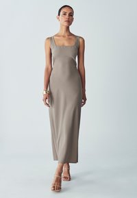 Langes, eng anliegendes Kleid aus glattem taupefarbenem Stoff mit einem quadratischen Ausschnitt, breiten Trägern und minimalistischem Design, kombiniert mit beigen Sandalen mit Riemchen.