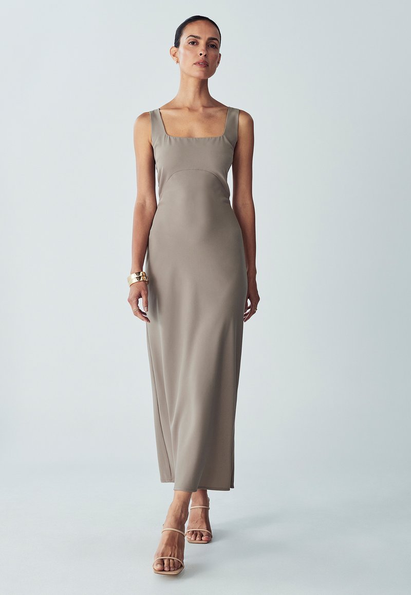 Langes, eng anliegendes Kleid aus glattem taupefarbenem Stoff mit einem quadratischen Ausschnitt, breiten Trägern und minimalistischem Design, kombiniert mit beigen Sandalen mit Riemchen.