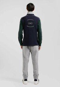 Pull en quarter-zip bleu marine et vert avec un col gris, arborant un patch logo au dos. Associé à un pantalon de jogging gris et des baskets.
