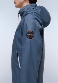 Waterafstotende jas in het blauw. Voorzien van een capuchon, trekkoord, zijzak met rits en een circulair logo op de mouw. Glad, strak textuur.