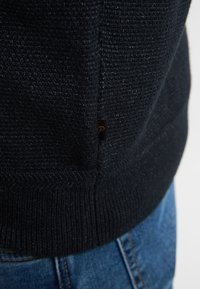 Maglione lavorato a maglia blu navy con una finitura testurizzata, dotato di orlo a coste e una piccola etichetta con logo sul lato. Adatto per un look casual.