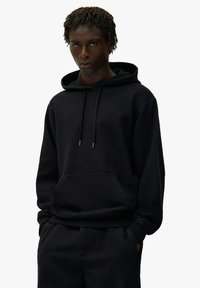 Mann trägt einen schwarzen Kapuzenpullover mit Kordeln, einer Kängurutasche und passenden schwarzen Hosen, steht mit den Händen in den Taschen.