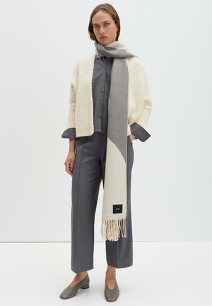 Cardigan crema lavorato a maglia indossato sopra una camicia grigia con bottoni, abbinato a pantaloni grigi e una sciarpa grigio chiaro con dettagli a frange. Scarpe verde oliva.