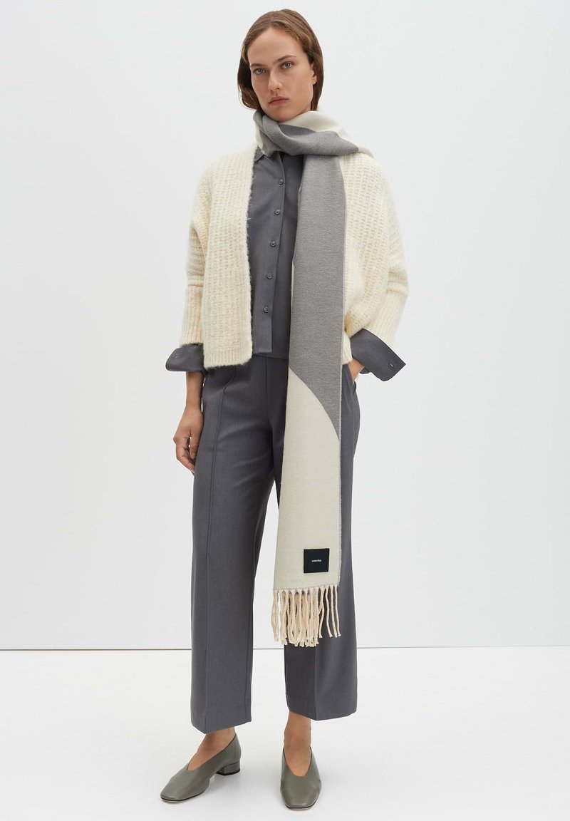 Gebreid crèmewit vest over een grijs overhemd met knopen, gecombineerd met een grijze broek en een lichtgrijze sjaal met franjes. Olijfgroene schoenen.