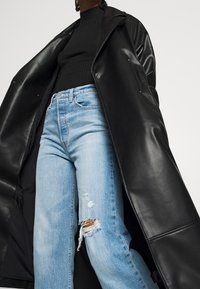 Long manteau en cuir noir porté sur un haut noir côtelé et un jean bleu clair déchiré avec ourlet effiloché et coutures visibles.