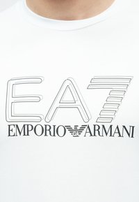 Κοντινό πλάνο λευκού πουκάμισου με το λογότυπο «EA7 Emporio Armani» σε μαύρο και γκρι γραμμές κατά μήκος του στήθους.
