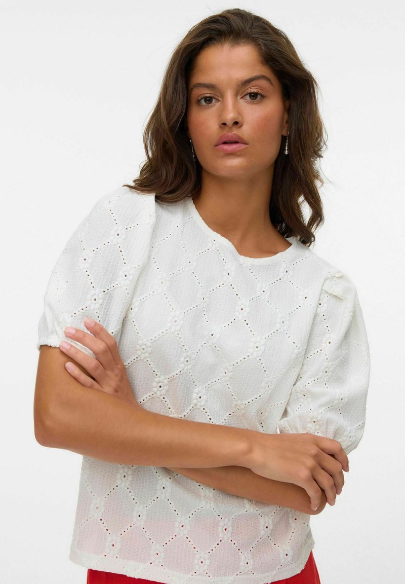 Hvit kortermet topp med et teksturert mønster som har utskårne diamanter og blomster, rund hals og løs passform.