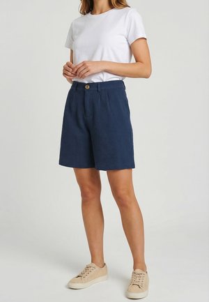 Donna che indossa una maglietta bianca a maniche corte, pantaloncini blu navy con trama e bottone, e sneaker beige con lacci, in piedi davanti a uno sfondo neutro.