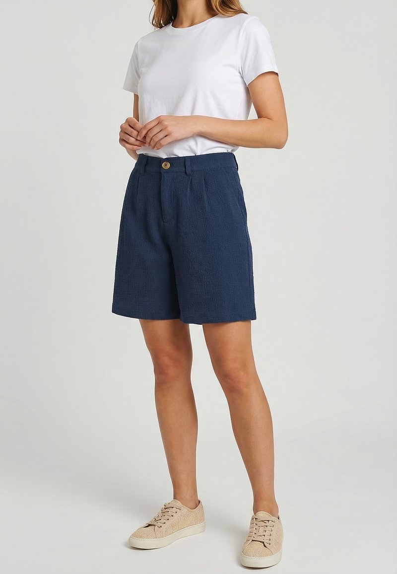 Mujer con camisa blanca de manga corta, pantalones cortos azul marino texturizados con botón y zapatillas beige con cordones, de pie frente a un fondo neutro.