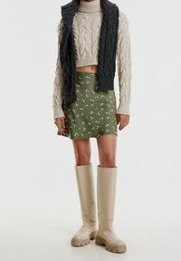 Lyhyeksi leikattu kermansävyinen palmikkoneule, vihreä kukkakuvioinen minihelma ja beige polvipituinen saappaat, joissa on kuvioitu pohja.