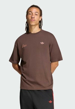 Uomo con dreadlocks che indossa una maglietta Adidas marrone con il testo "Paris" e pantaloni neri con strisce rosse e logo Adidas, in piedi con le mani dietro la schiena.