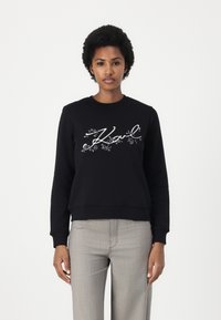 Sudadera negra con cuello redondo, con estampados florales blancos y la palabra "Karl" en cursiva en el frente. Tejido de mezcla de algodón.