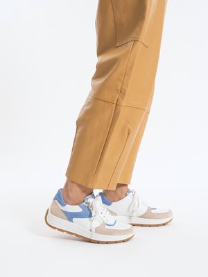 Person wearing tan cropped pants and white sneakers with blue and beige suede accents on a white background.
