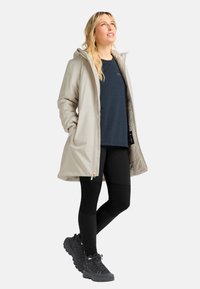 Frau steht mit den Händen in den Taschen, trägt einen beigen Kapuzenmantel, ein navyfarbenes Shirt, schwarze Leggings und schwarze Wanderschuhe und schaut nach oben.