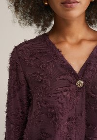 Texturerad mörklila blus med V-ringning och framknäppning med en gyllene blommönster; tyget har intrikata lager av fransar.