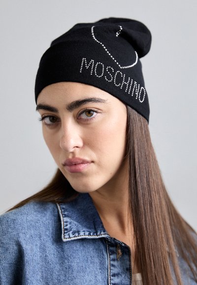 Jonge vrouw met lang steil haar draagt een zwarte beanie met de tekst "Moschino" en een denim jasje tegen een effen lichte achtergrond.