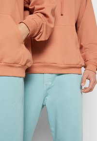 Dwie osoby stoją obok siebie, ubrane w brzoskwiniowe bluzy z kapturem i jasnoniebieskie dżinsy, jedna z rękami w kieszeni bluzy, a druga z luzem opuszczonymi rękami.