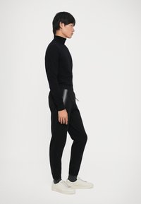 Jovem vestido com um suéter de gola alta preto, calças de jogging pretas com detalhes em zíper, meias escuras e ténis brancos, posicionado de lado sobre um fundo branco.